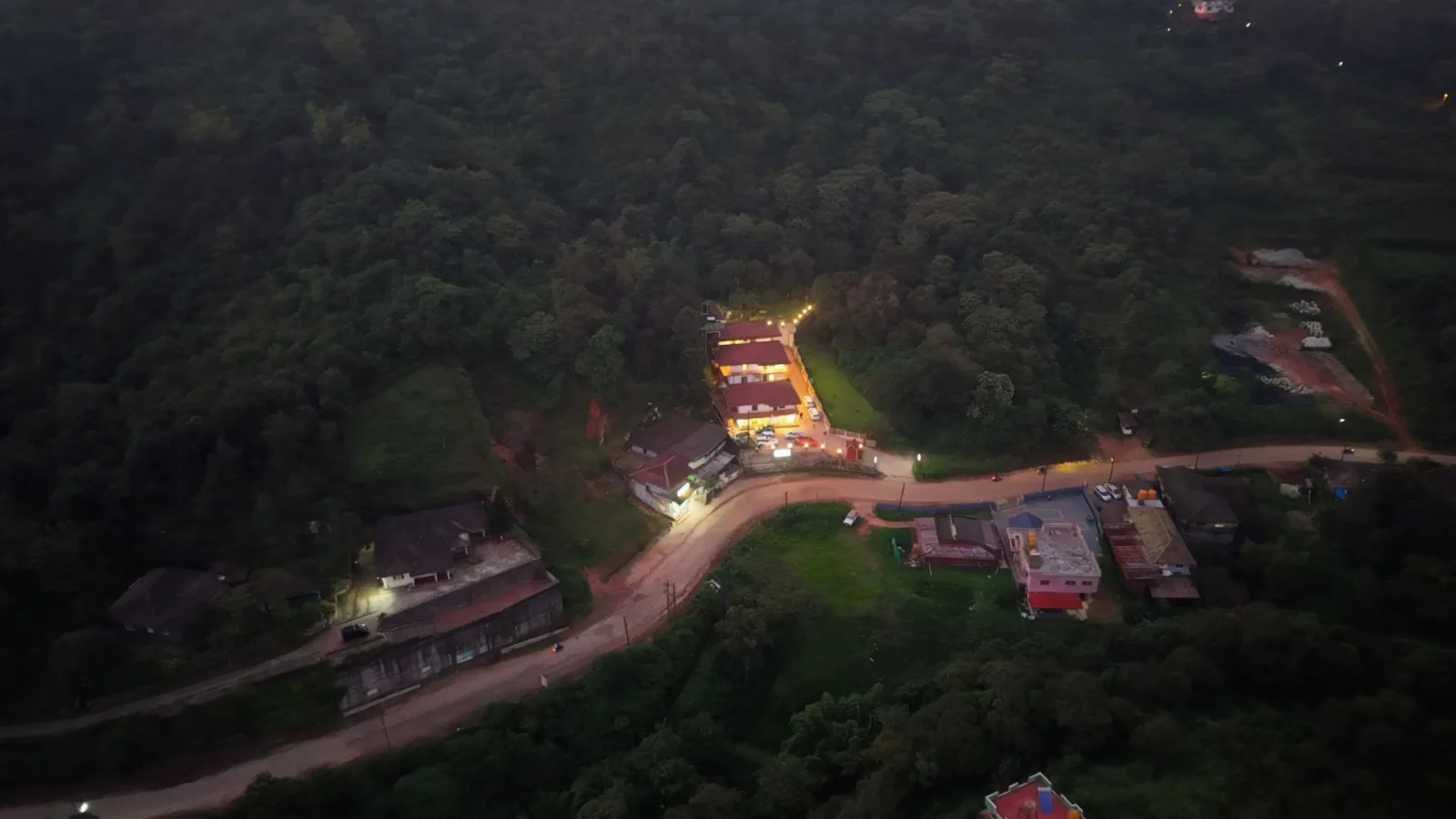 Hasu Stay seen from the air at dusk, a small lit property surrounded by forested hills outside Madikeri